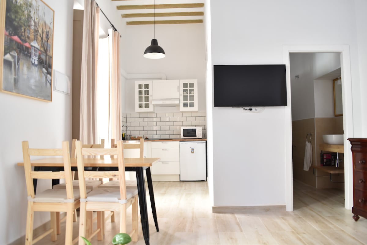 An open kitchen and dining area is presented, featuring a wooden table with four chairs. The kitchen includes white cabinetry, a microwave, and a refrigerator. A wall-mounted TV is visible, along with a doorway leading to a bathroom. Soft natural light illuminates the space.