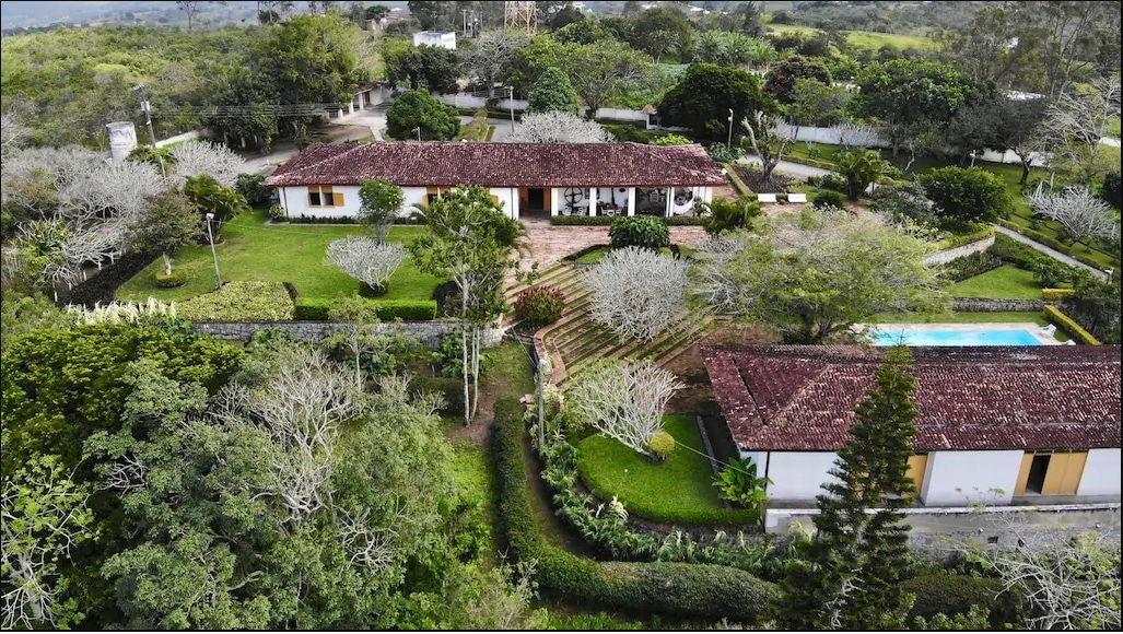 An aerial view captures the expansive private property surrounded by lush greenery and gardens. The single-story house features a red-tiled roof and is complemented by a swimming pool, with well-maintained pathways and trees offering a serene atmosphere.