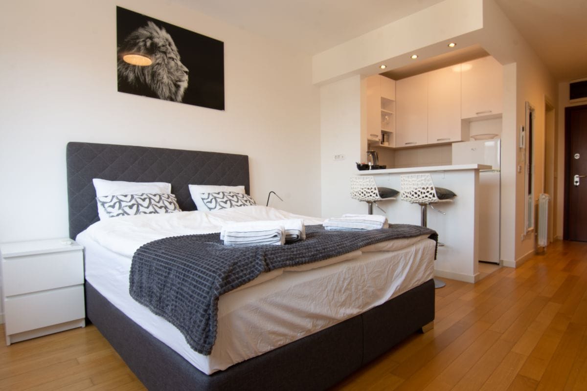 A spacious bedroom is furnished with a large bed topped with two sets of pillows. A cozy throw blanket is draped across the bed. A black and white lion portrait adorns the wall above. A bar-style table with two stools is positioned in the background.