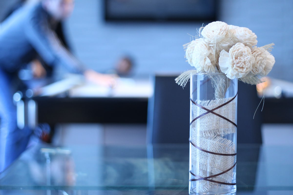 A glass vase filled with light-colored, dried flowers is positioned on a dining table. The blurred background features people engaged in a game, indicating a lively, communal atmosphere.