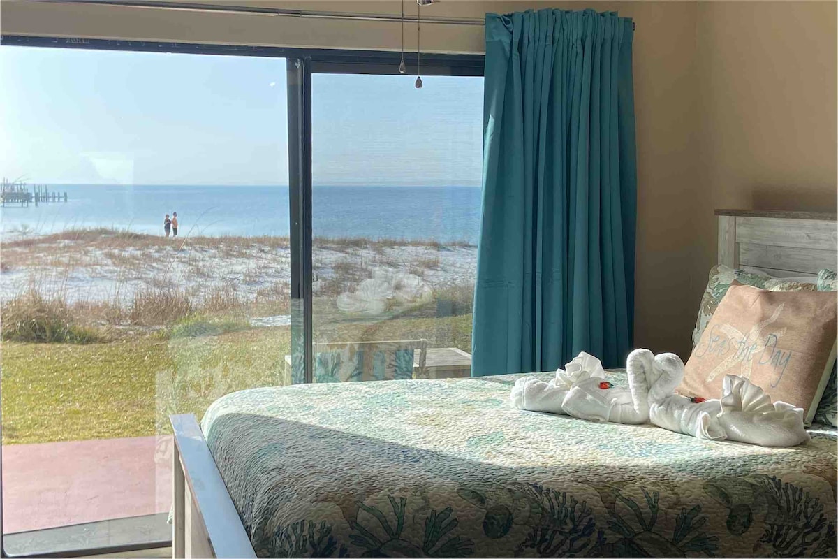 A spacious bed is adorned with a light teal quilt and neatly arranged plush pillows. Large glass doors offer a clear view of the waterfront, with gentle waves and a distant fishing pier visible. Natural light fills the room through the sheer curtains.