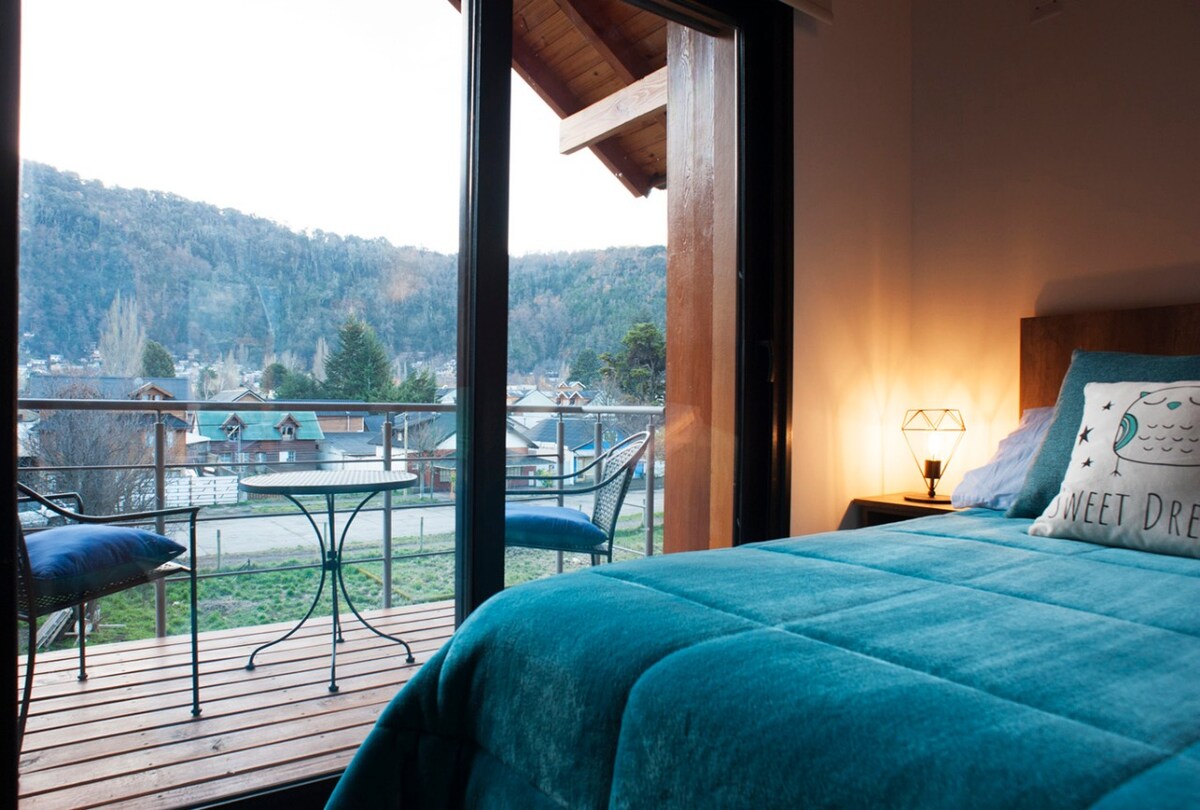 A cozy sleeping area is visible, featuring a spacious bed dressed in a soft blue coverlet. A balcony is accessed through large glass doors, showcasing a view of the mountains and nearby residential area. A small table and two chairs are placed outside.