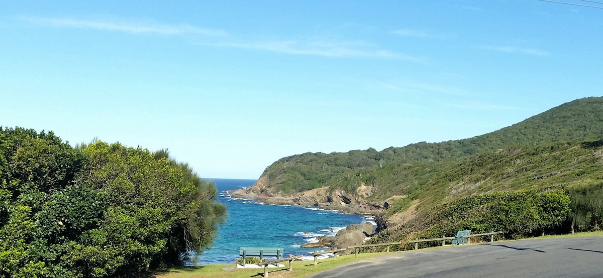 A scenic coastal view features a rocky shoreline meeting the clear blue ocean. Lush greenery lines the foreground, with gentle hills in the background. A narrow road curves along the coast, providing a tranquil atmosphere and inviting natural surroundings.