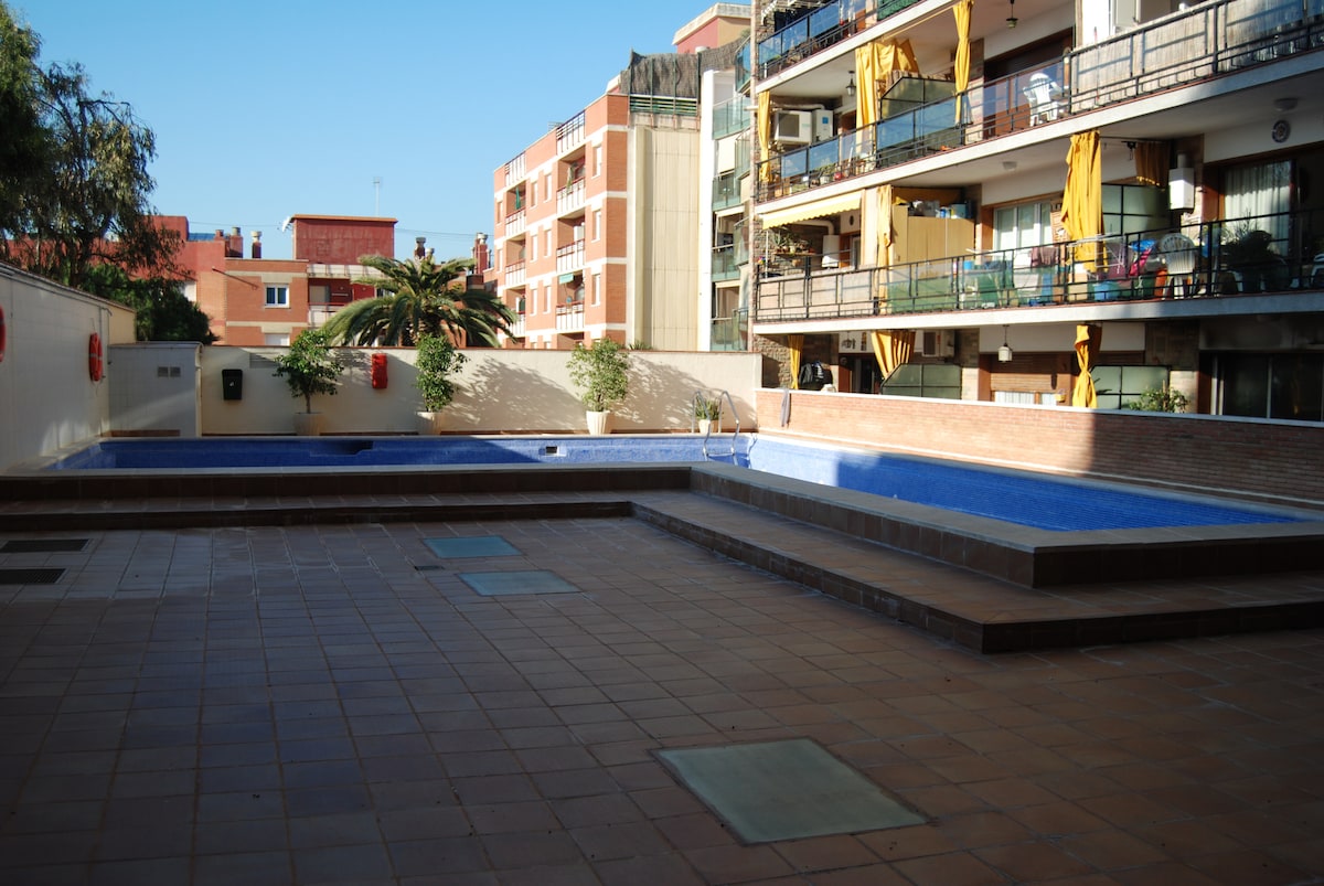 An outdoor pool area is presented, featuring a rectangular pool surrounded by tiled surfaces. Apartment balconies overlook the scene, with greenery partially visible. Sunlight illuminates the area, indicating a clean and well-maintained environment, suitable for relaxation.