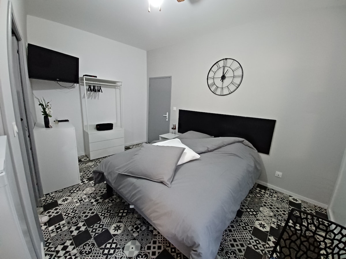 A spacious sleeping area features a large bed with gray bedding and decorative pillows. A wall-mounted television is positioned nearby, and a stylish clock is displayed above the headboard. The floor showcases a patterned design, contributing to the room's contemporary aesthetic.