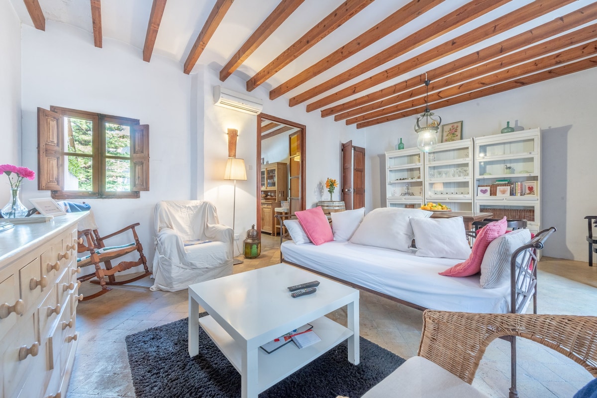 The living area features a white sofa adorned with pink cushions, complemented by a rocking chair and a wicker armchair. Exposed wooden beams line the ceiling, while large windows allow natural light to fill the space. A modern coffee table sits atop a textured black rug.