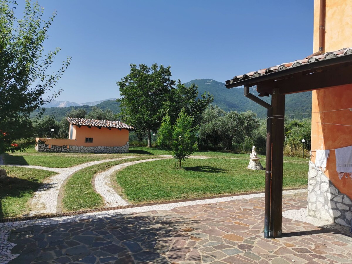 A well-maintained outdoor garden space features lush greenery and winding pathways. A small building is located in the background, surrounded by trees. The mountains create a scenic backdrop, enhancing the natural setting.