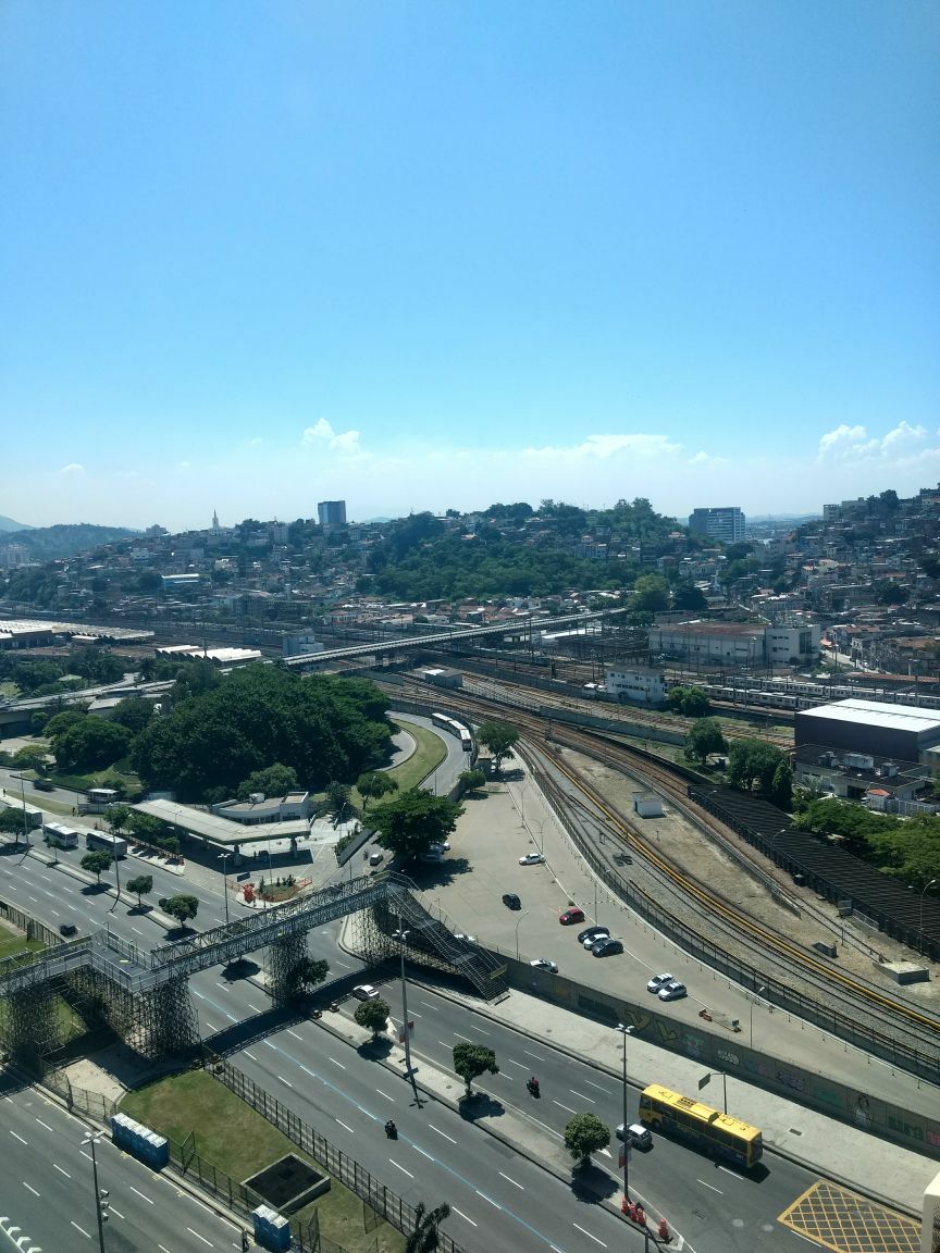 A view from the property displays a broad cityscape, featuring a network of roads and railways. Lush greenery is interspersed with urban structures, while a distant hillside is visible under a clear blue sky dotted with clouds. Urban development and transportation infrastructure are prominent.