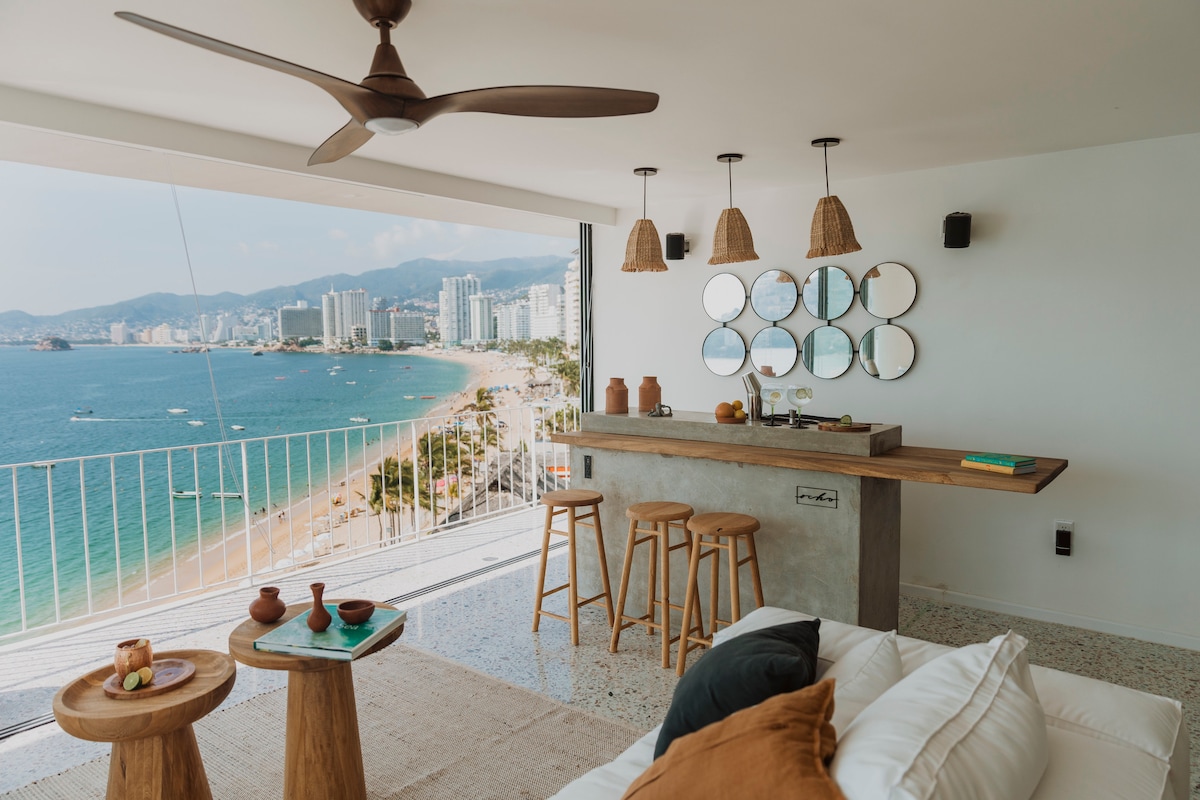 A spacious living area features large floor-to-ceiling windows showcasing panoramic views of the beach and bay. A sleek bar with three wooden stools provides additional seating. Natural light illuminates the space, while a ceiling fan offers comfort. Decorative mirrors and soft furnishings add a cozy touch.