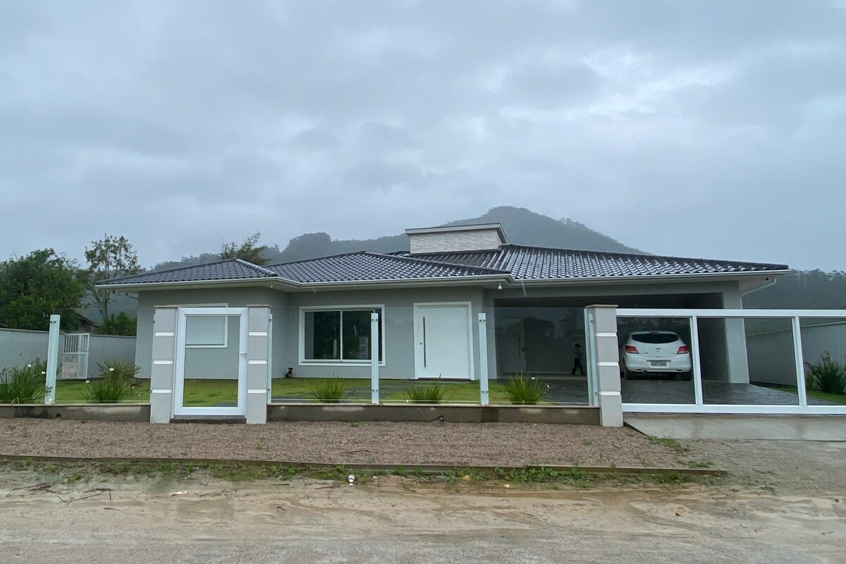 The modern house features a sleek design with a metal roof and large windows, allowing natural light to enter. A car is parked in the covered area, and the front yard is lined with greenery and gravel.