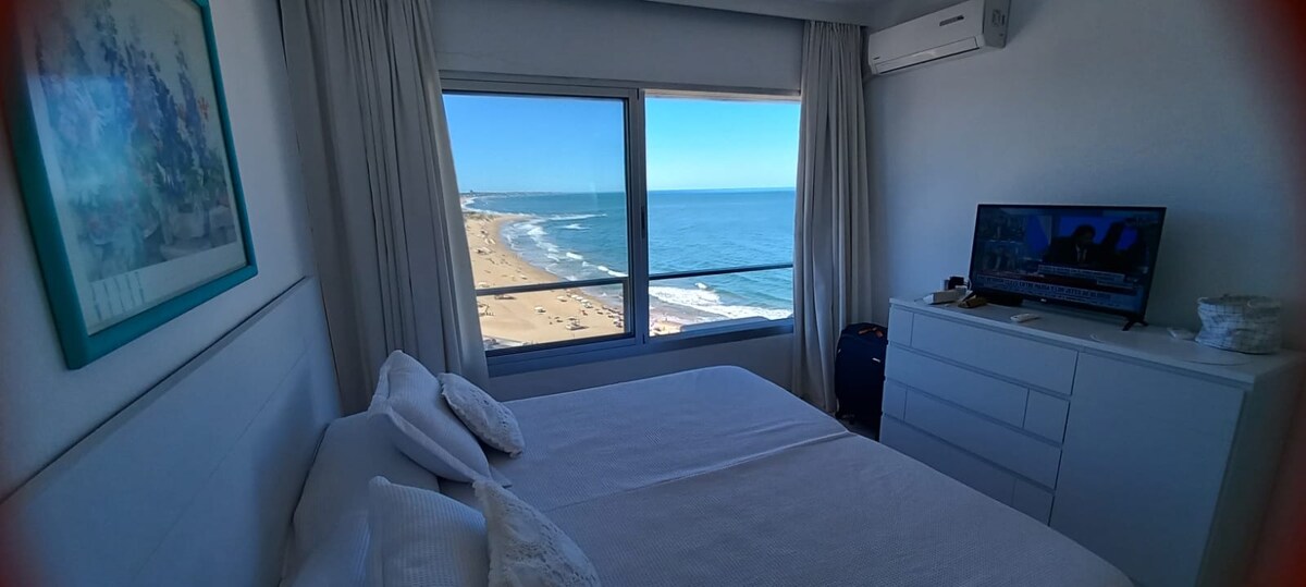 A well-lit bedroom features a spacious bed with soft white bedding. Large windows offer panoramic views of the coastline and ocean, while a dresser and television complete the room's amenities. Light curtains frame the window, enhancing the bright atmosphere of the space.