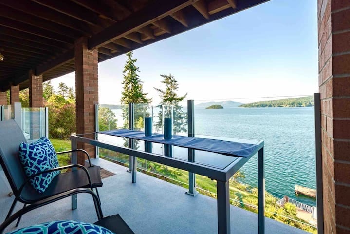 A spacious patio is shown with a dining table set for outdoor meals. Comfortable chairs are positioned alongside, all overlooking the tranquil ocean waters. Lush vegetation and distant islands can be seen in the backdrop, providing a serene and relaxing ambiance.