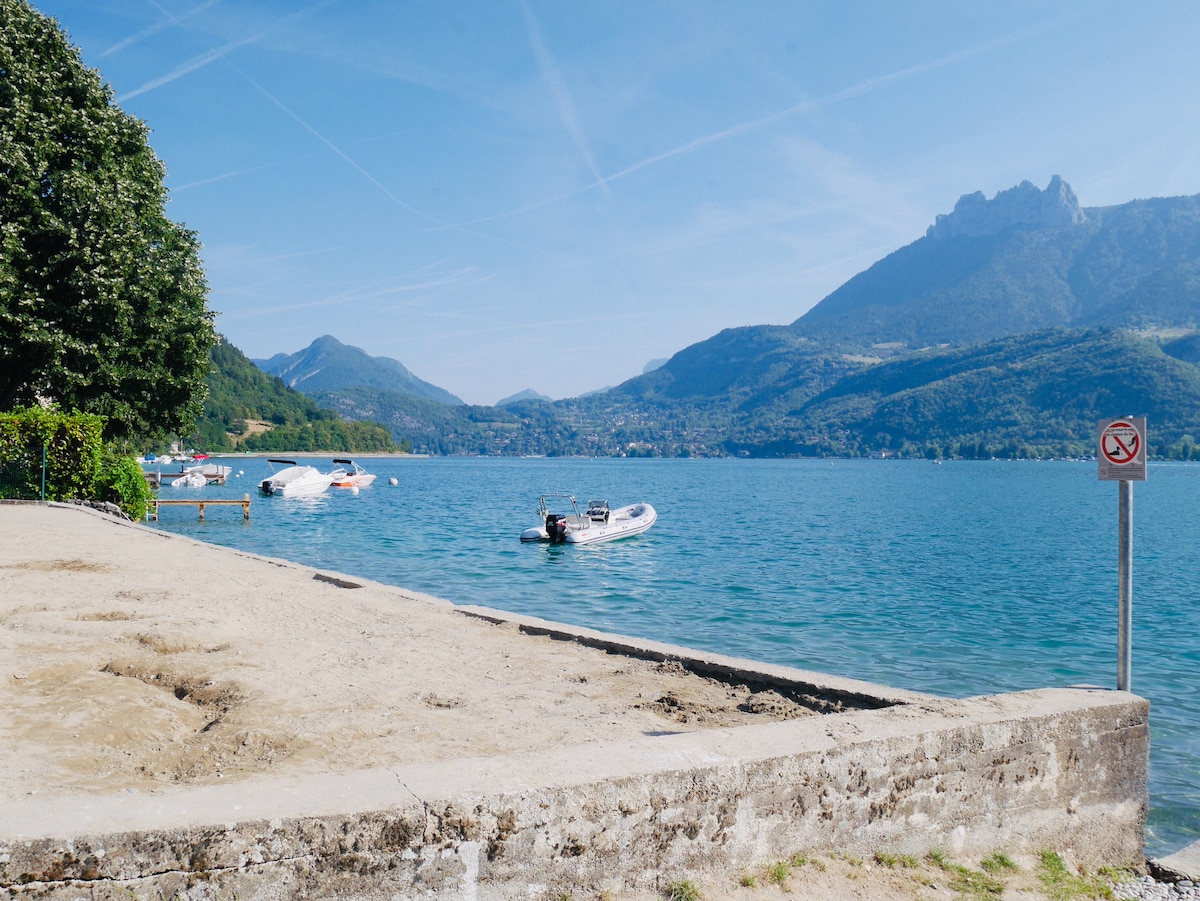 A sandy area borders the serene waters of Lake Annecy, where several boats are anchored. In the background, lush green hills rise against a clear blue sky, while a mountainous silhouette completes the scenic view. A no swimming sign is placed near the edge of the sandy shore.