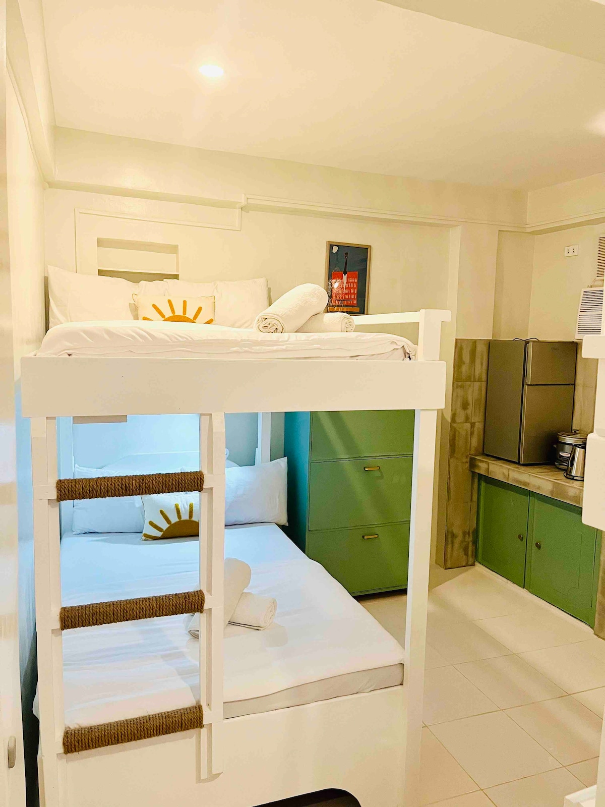 The image displays a room featuring two double deck beds, accommodating a total of six guests. A small kitchenette is visible, equipped with a refrigerator and coffee maker. Clean linens and towels are neatly arranged on the beds, contributing to a tidy and organized ambiance.