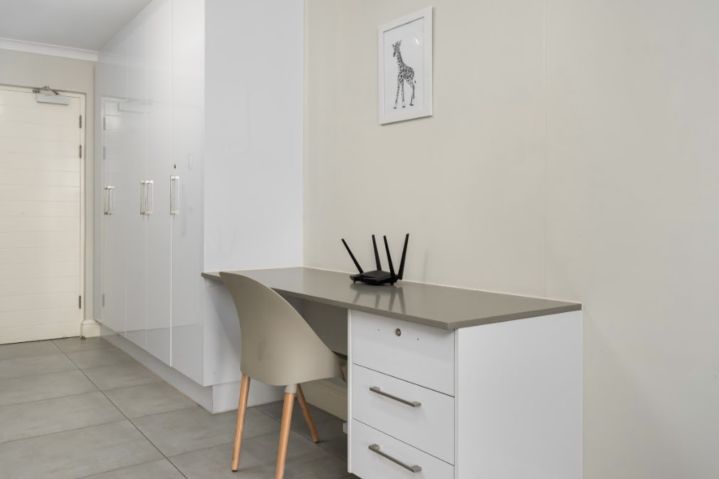 A modern workspace is depicted, featuring a sleek desk with a smooth surface and a drawer unit. A stylish chair complements the desk, while a router is positioned on the tabletop. A framed giraffe illustration is hung above the desk, adding a light touch to the area.