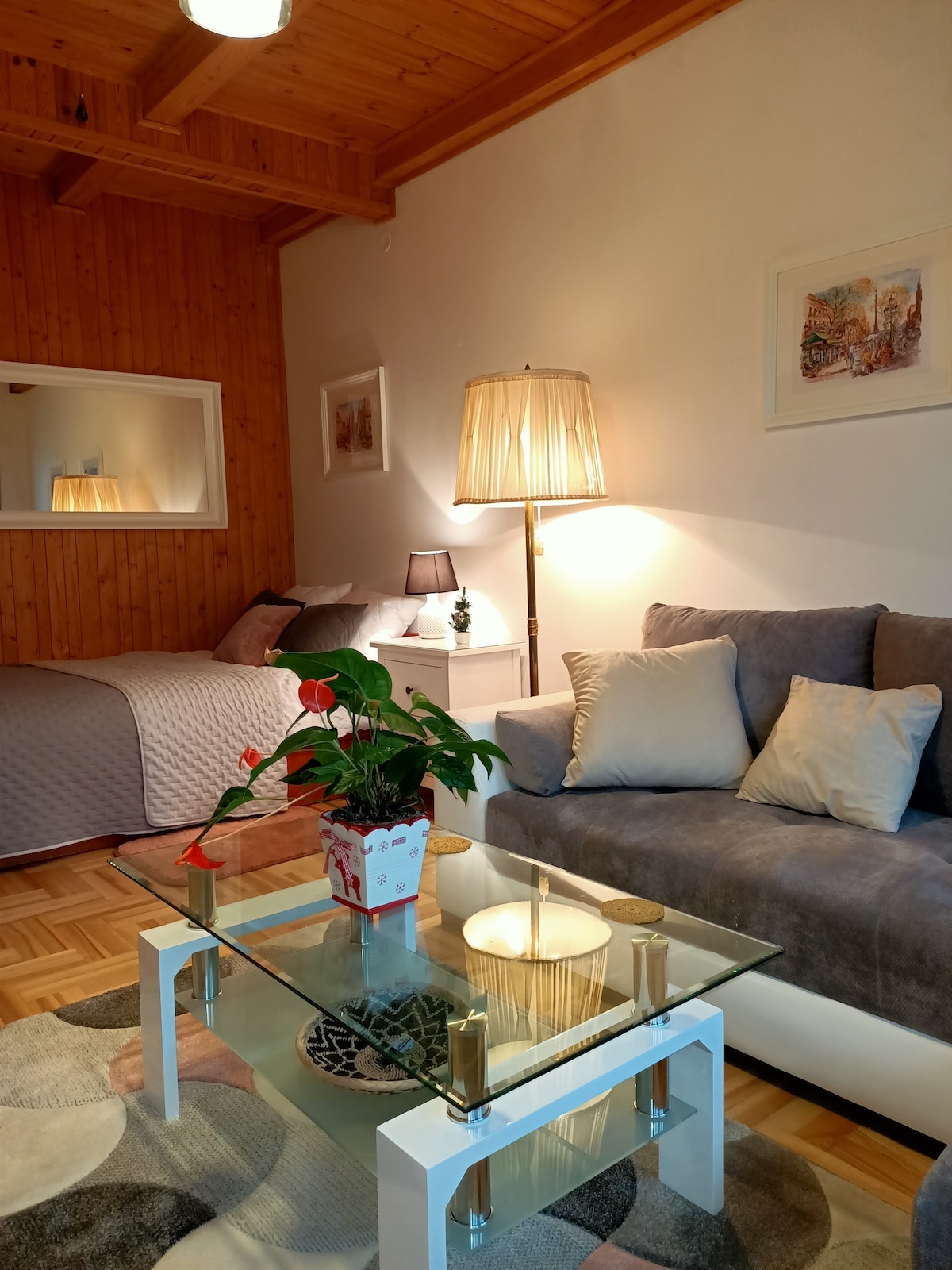 A cozy living area features a comfortable sofa and a glass coffee table, surrounded by soft decorative pillows. A small potted plant adds a touch of greenery. In the background, a well-made bed is visible, complemented by warm wooden accents and soft lighting.