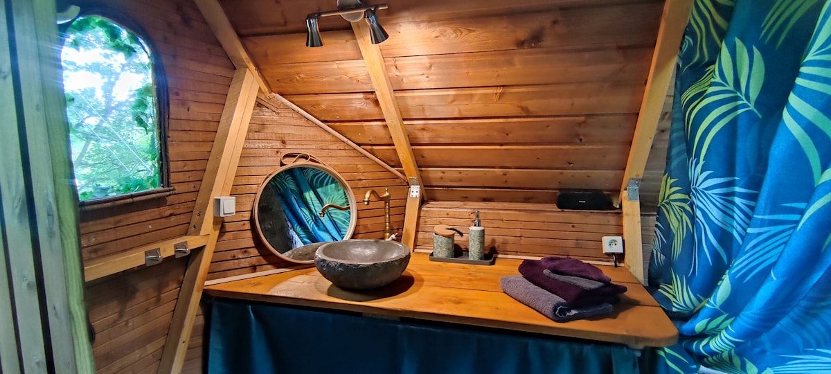 A rustic wooden vanity is featured, showcasing a round mirror and a natural stone sink. Towels are neatly arranged beside a collection of toiletries. The interior walls, adorned with wood paneling, create a warm atmosphere, while a tropical-patterned curtain adds a splash of color.