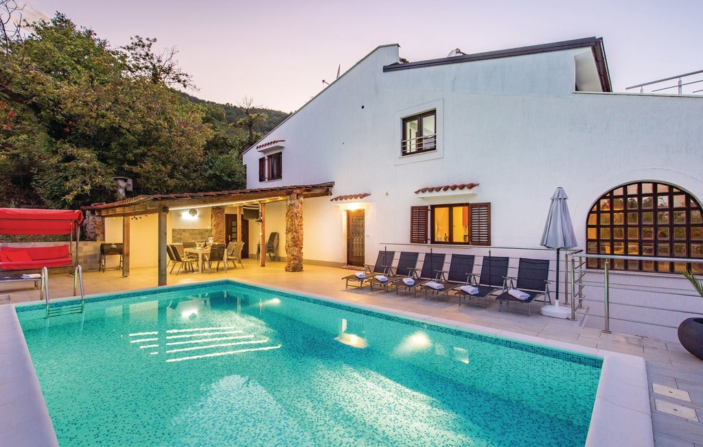 A refreshing outdoor pool with turquoise water is surrounded by sun loungers. A shaded patio area with dining furniture is visible, along with a charming stone structure. The exterior features a blend of white walls and wooden accents, complemented by greenery in the background.