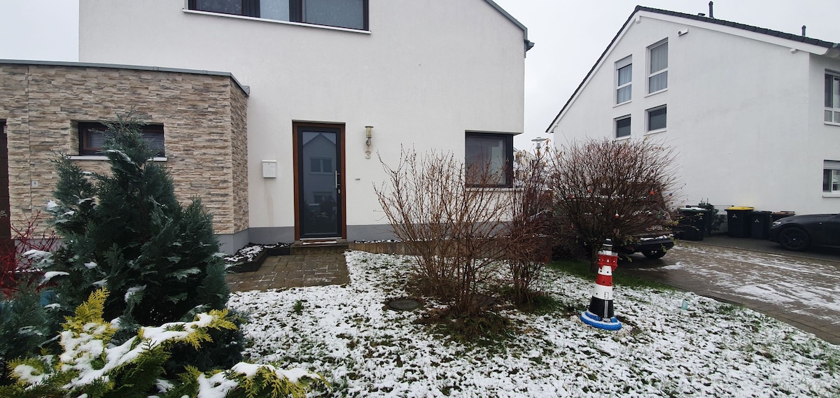 The exterior of the apartment is displayed, featuring a simple entrance surrounded by a small landscaped area. Snow covers parts of the ground, while shrubs and plants add greenery. Nearby houses are visible, highlighting the quiet residential setting.