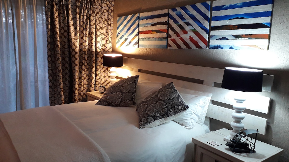 A modern bedroom features a spacious bed with white linens and decorative pillows. Two bedside lamps provide soft lighting, complementing the colorful abstract art above the headboard. Light filters through sheer curtains, creating a soothing ambiance in the room.