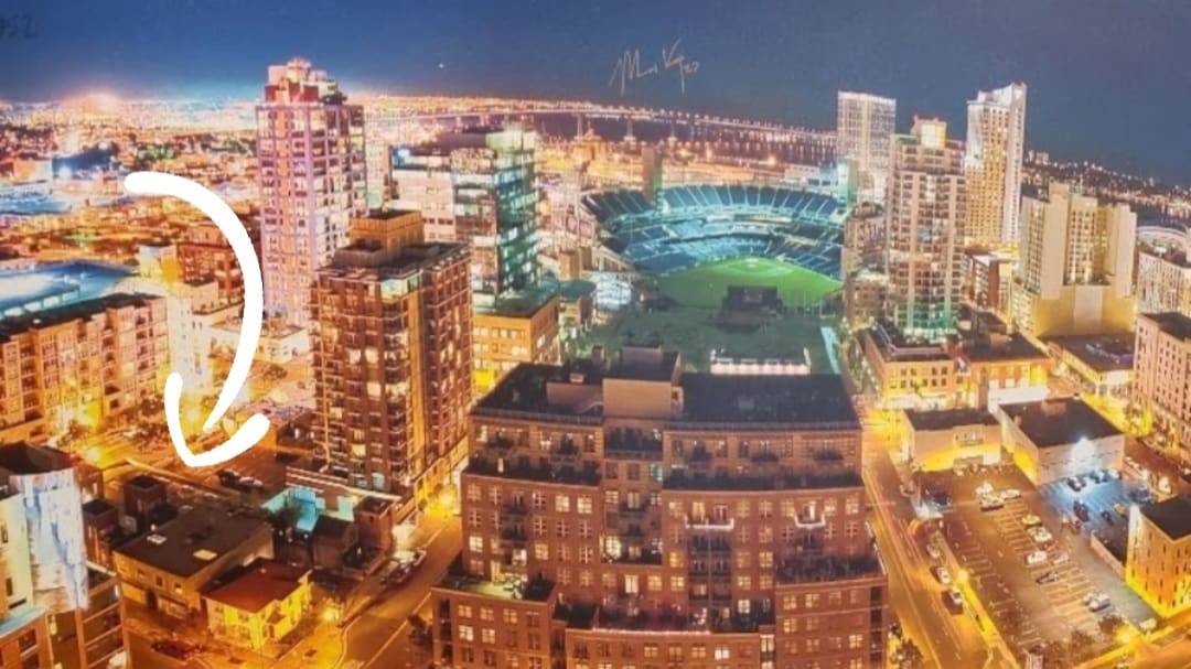 An aerial view of downtown San Diego at night, showcasing the illuminated skyline and bustling streets. The bright lights of buildings contrast with the darkness of the night sky. A ballpark is visible, providing insight into the urban environment.