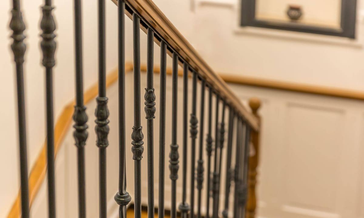 A close-up view of an ornate staircase railing is presented, featuring dark metal balusters with decorative elements. The wood of the handrail offers a contrasting warm tone. The surrounding walls are painted in a light color, enhancing the spacious feel of the staircase area.