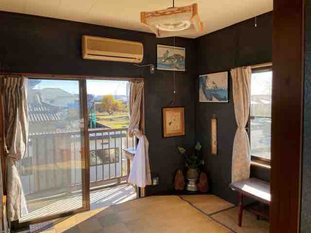 A cozy room features tatami flooring and large windows allowing natural light. Air conditioning is visible on the wall, while decorative artwork adorns the dark walls. Sheer curtains frame the window, offering a view of the outdoors. A seating area is positioned next to the window.