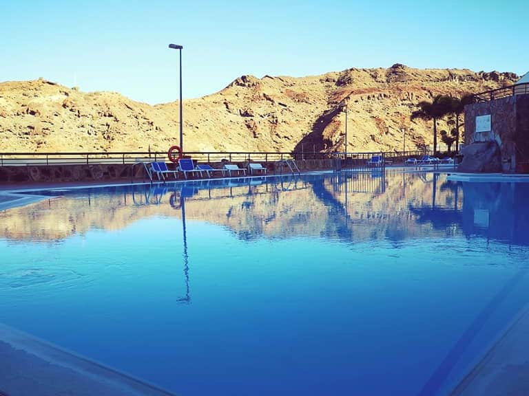 A large swimming pool is visible, reflecting the clear blue sky and surrounding mountainous landscape. Sun loungers are arranged neatly along the poolside, offering a space for relaxation. The tranquil water invites refreshing leisure with scenic views of the rugged terrain.