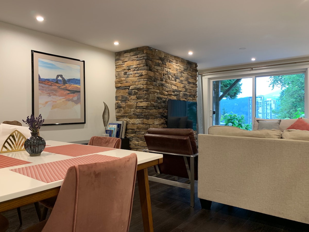 The living area presents a modern design with a stone wall feature. A dining table is set with a checkered placemat, surrounded by upholstered chairs. Large windows allow natural light to fill the space, offering a view of the outdoors and enhancing the open feel.