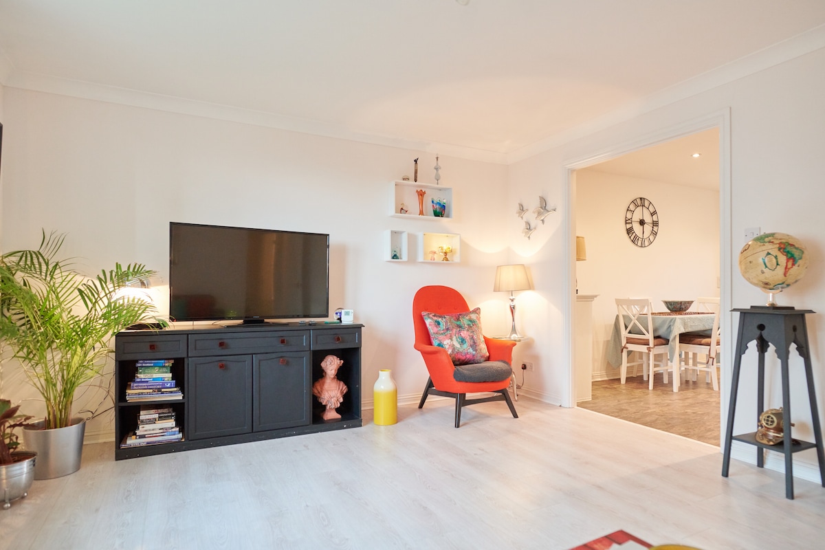 A cozy living area features a flat-screen television atop a black cabinet. A vibrant orange chair adds a pop of color, complemented by a floor lamp. To the side, a leafy plant and decorative shelves with items create a comfortable atmosphere. A doorway leads to the dining area.