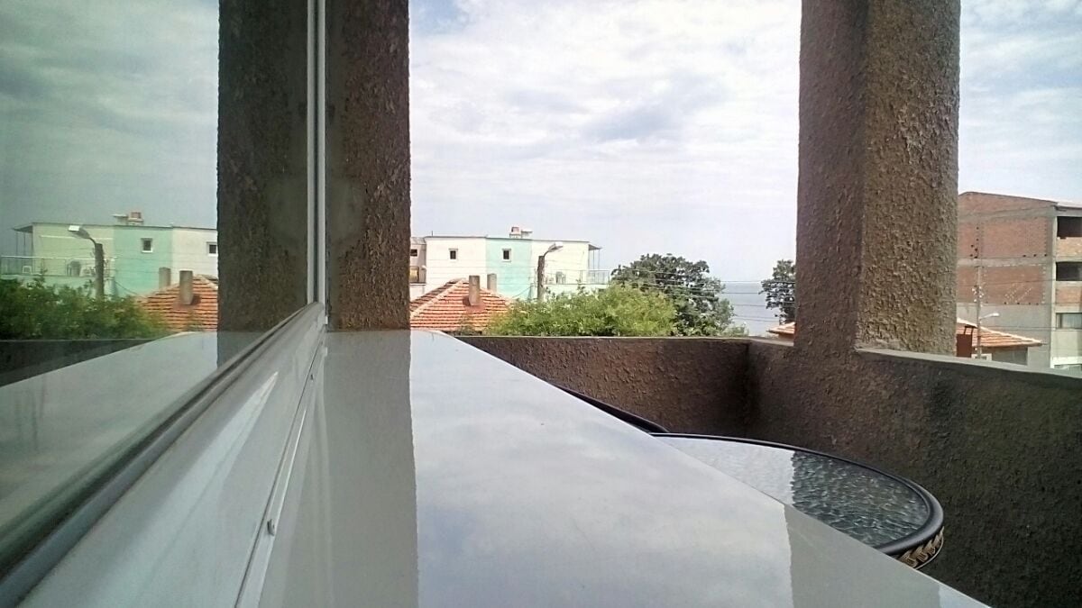 A view from a balcony is visible, showcasing the horizon where buildings meet the sea. A round table with a glass top is perched on a smooth ledge, reflecting natural light. The surrounding greenery adds a touch of nature to the urban landscape.