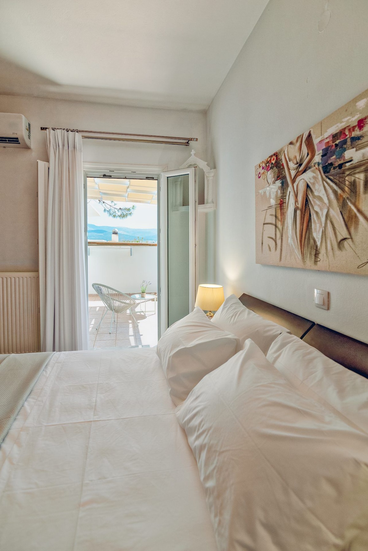 A serene bedroom features a spacious bed with crisp white bedding and plush pillows. Natural light enters through an open door, leading to a cozy balcony adorned with a chair. A piece of artwork hangs on the wall, adding a touch of character to the space.