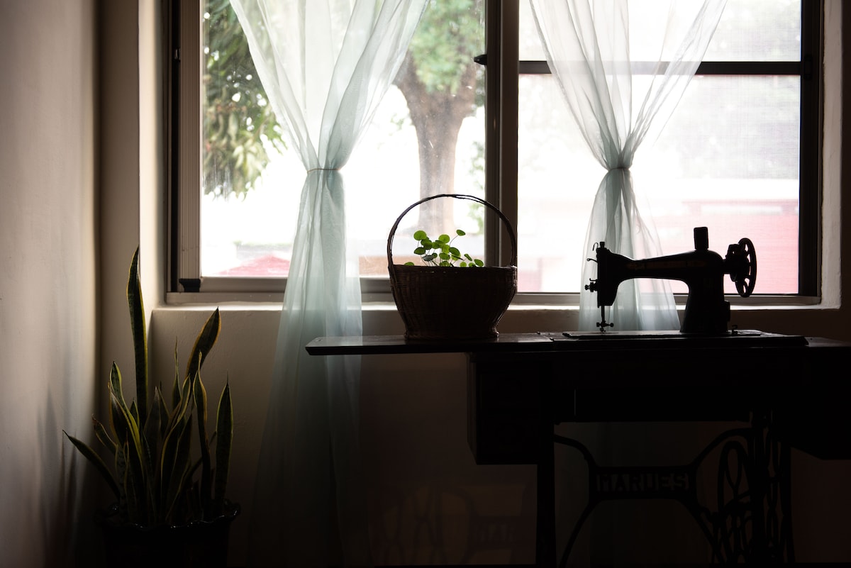A window draped with light, sheer curtains allows natural light to filter in, illuminating a small potted plant in a woven basket. Beside it, an antique sewing machine adds a touch of vintage charm to the room. A tall plant is visible on the floor.