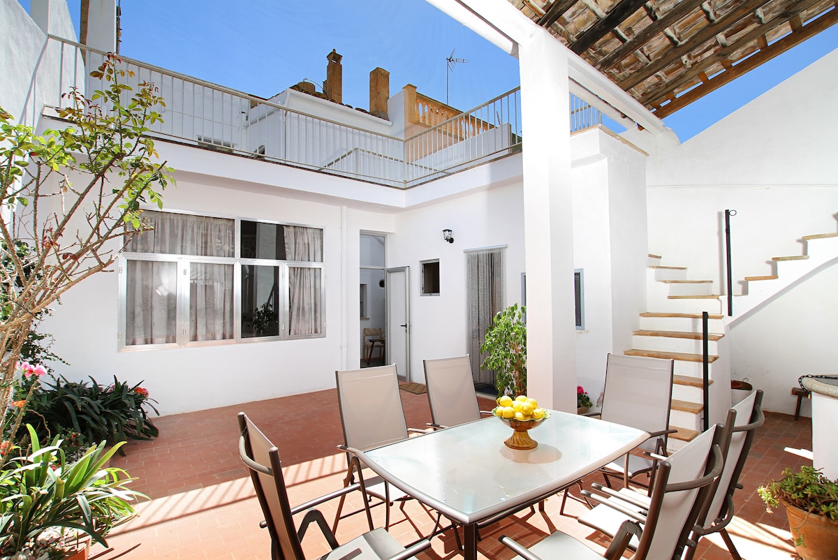 An enclosed terrace area is presented, featuring a large table set for dining with several chairs surrounding it. Lush greenery adorns the corners, while a staircase leads to an upper level. Bright light filters in through the nearby windows.