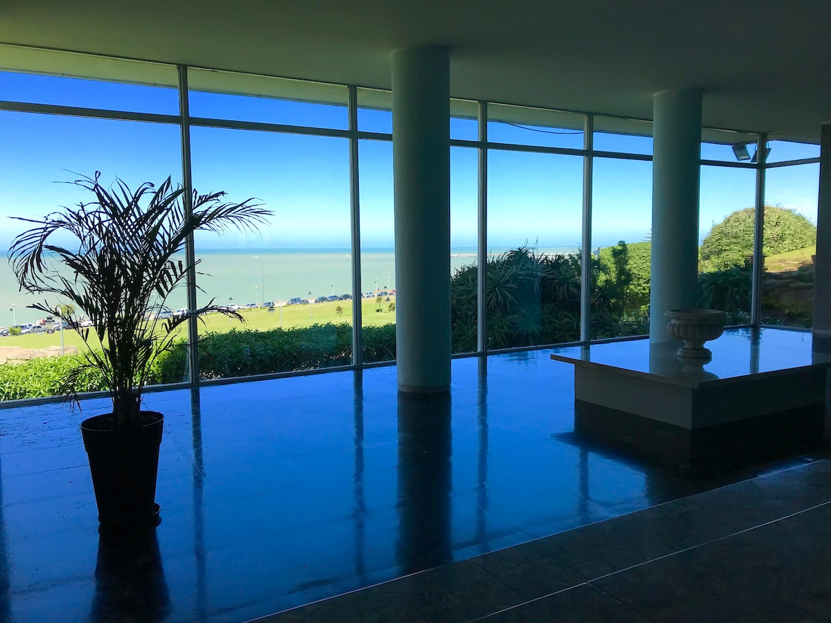 A bright communal area is illuminated by large glass windows offering expansive views of the sea and surrounding greenery. A tall potted palm plant adds a touch of nature, while the polished floor reflects the light and creates a spacious feel.