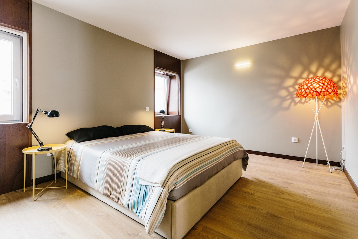 A spacious sleeping area features a large bed with neutral bedding and subtle striped accents. Natural light enters through two windows, enhancing the earthy tones of the walls. A contemporary floor lamp and a round side table add functional elements to the room's design.
