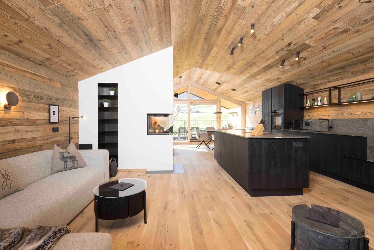 The living area features warm wooden walls and flooring, creating a natural ambiance. A modern kitchen is visible with dark cabinetry, an island, and light wood accents. A cozy sofa and round coffee table invite relaxation, with large windows allowing natural light to fill the space.