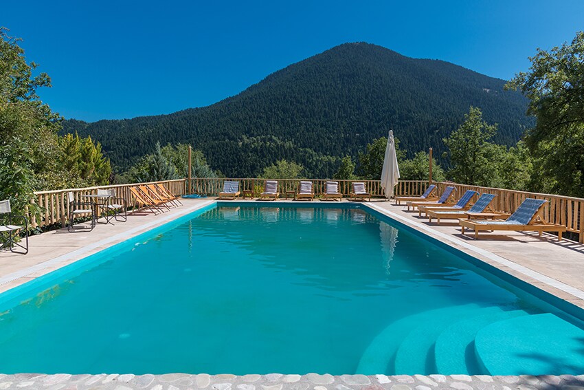 A spacious outdoor swimming pool is bordered by wooden decking, showcasing multiple sun loungers arranged for relaxation. The serene water reflects the clear blue sky, while lush greenery and a distant mountain backdrop enhance the tranquil setting.