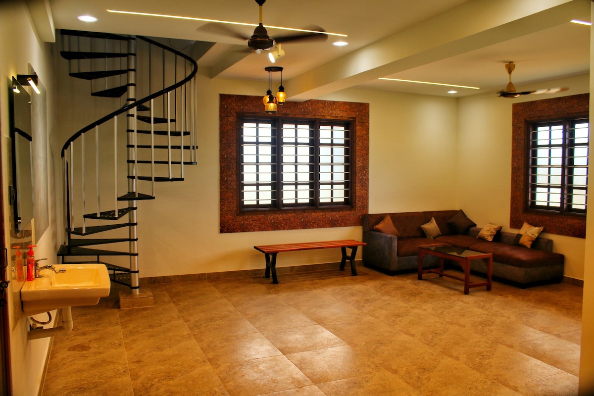 A spacious living area is highlighted by a cozy arrangement of a dark sofa and a wooden coffee table. A staircase with a spiral design leads to the upper level. Large windows provide ample natural light, complemented by soft overhead lighting.