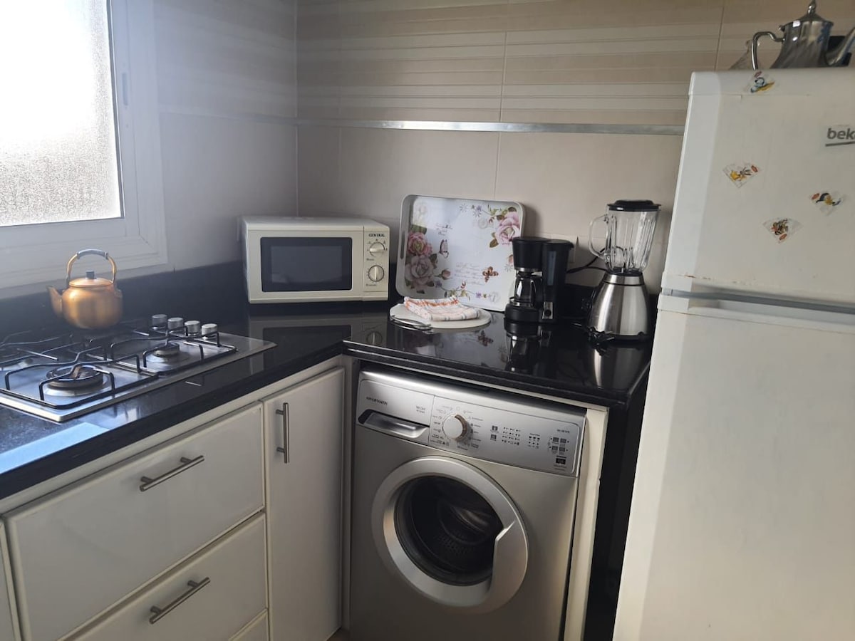 The kitchen is equipped with modern appliances including a washing machine, microwave, and coffee maker. A gas stove is present alongside a sleek black countertop. A decorative tray with floral design adds a touch of charm to the space. Natural light filters in through a nearby window.