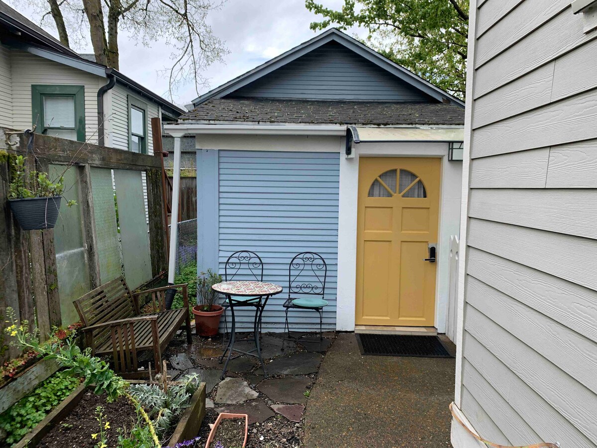 An inviting outdoor area leads to the bright yellow door of The Hen House. A small circular table with two chairs is positioned on a stone path, surrounded by garden beds with various plants, providing a peaceful entrance to the converted garage.
