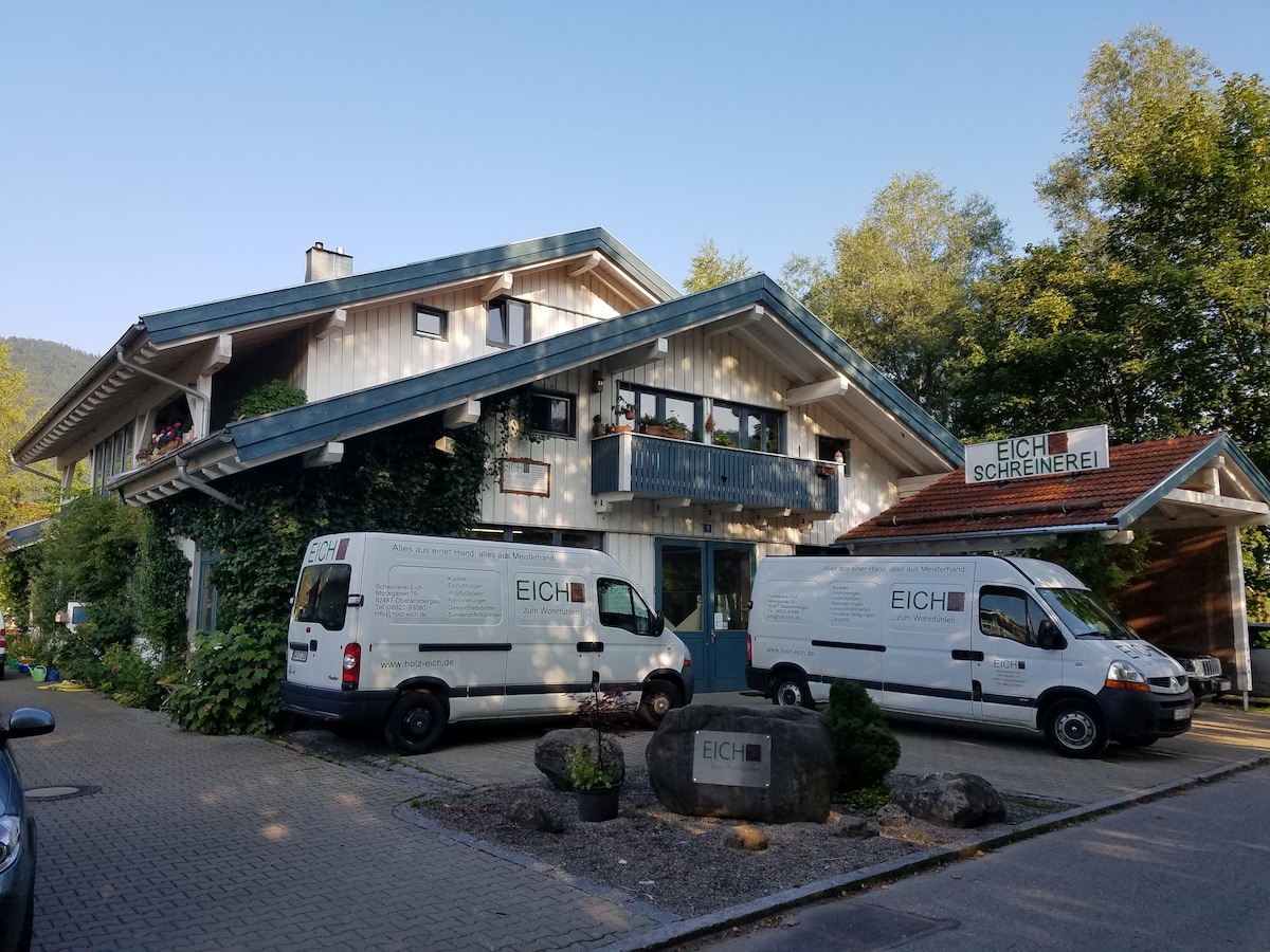 The exterior of a multi-story building is shown, featuring a triangular roof and multiple balconies. Two vans are parked in front, labeled with the name 'EICH'. Lush greenery surrounds the building, enhancing its natural setting.