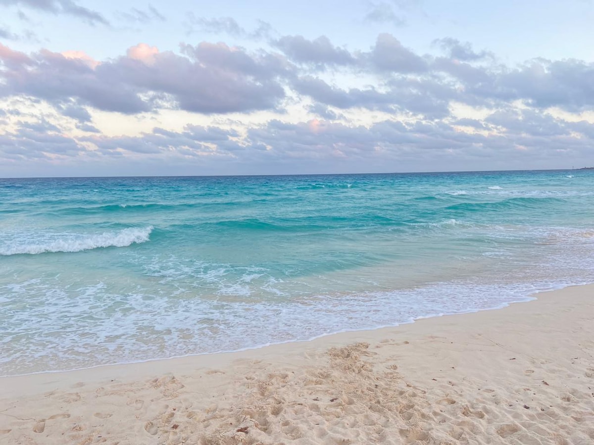 Soft waves lap gently against a sandy beach, with the ocean transitioning from turquoise to deep blue. Fluffy clouds scatter across a soft pastel sky, reflecting the tranquil atmosphere of a beachfront view.
