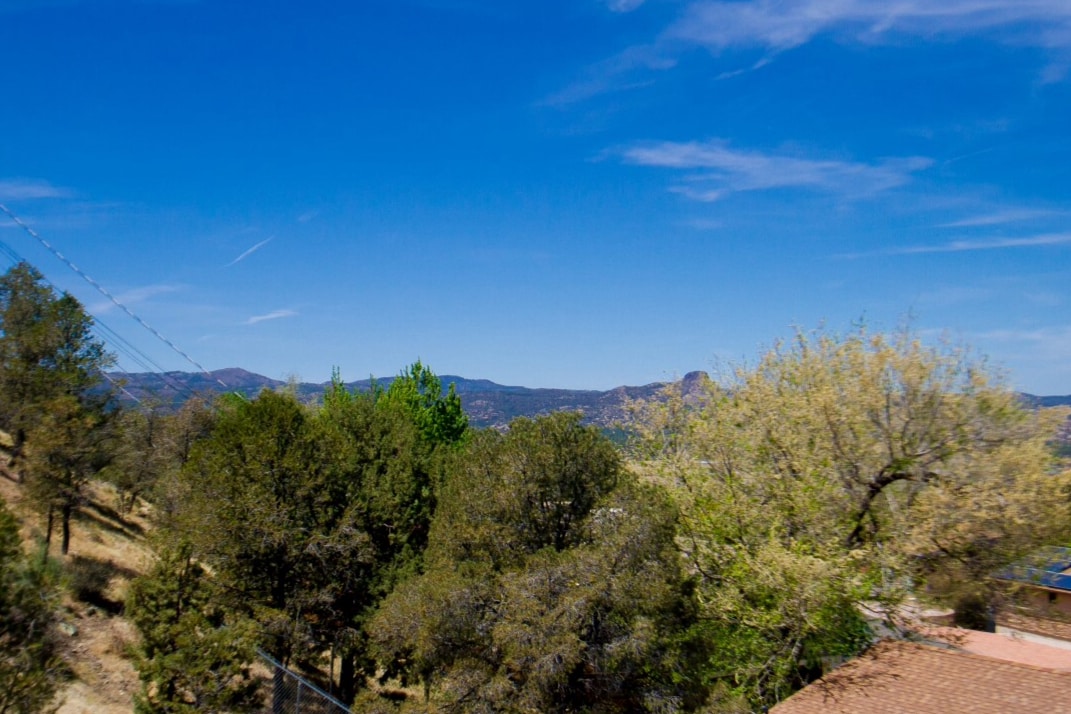A panoramic view showcases rolling hills and lush greenery under a clear blue sky. Trees dot the landscape, providing depth to the scenery, while distant mountains can be seen on the horizon.