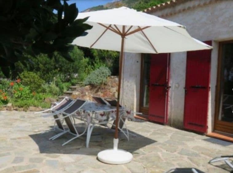 An outdoor terrace features a large white umbrella providing shade over a table surrounded by six chairs. The stone flooring adds a rustic charm, while red double doors lead into the dwelling. Lush greenery and colorful flowers enhance the natural setting.