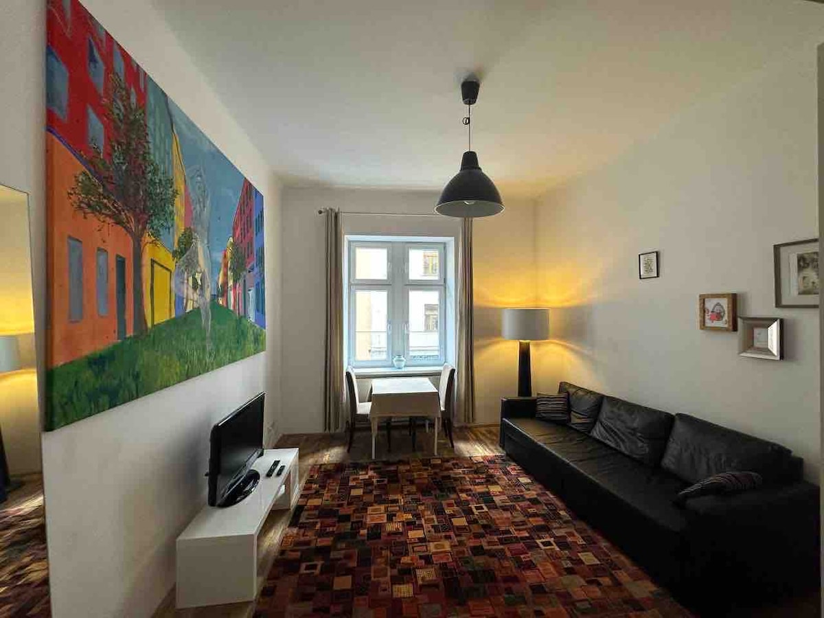 A spacious living area features a large black sofa positioned beside a modern TV stand. A vibrant painting adorns one wall, while the opposite wall includes a window with soft natural light. A dining table is set for four, illuminated by a ceiling lamp.