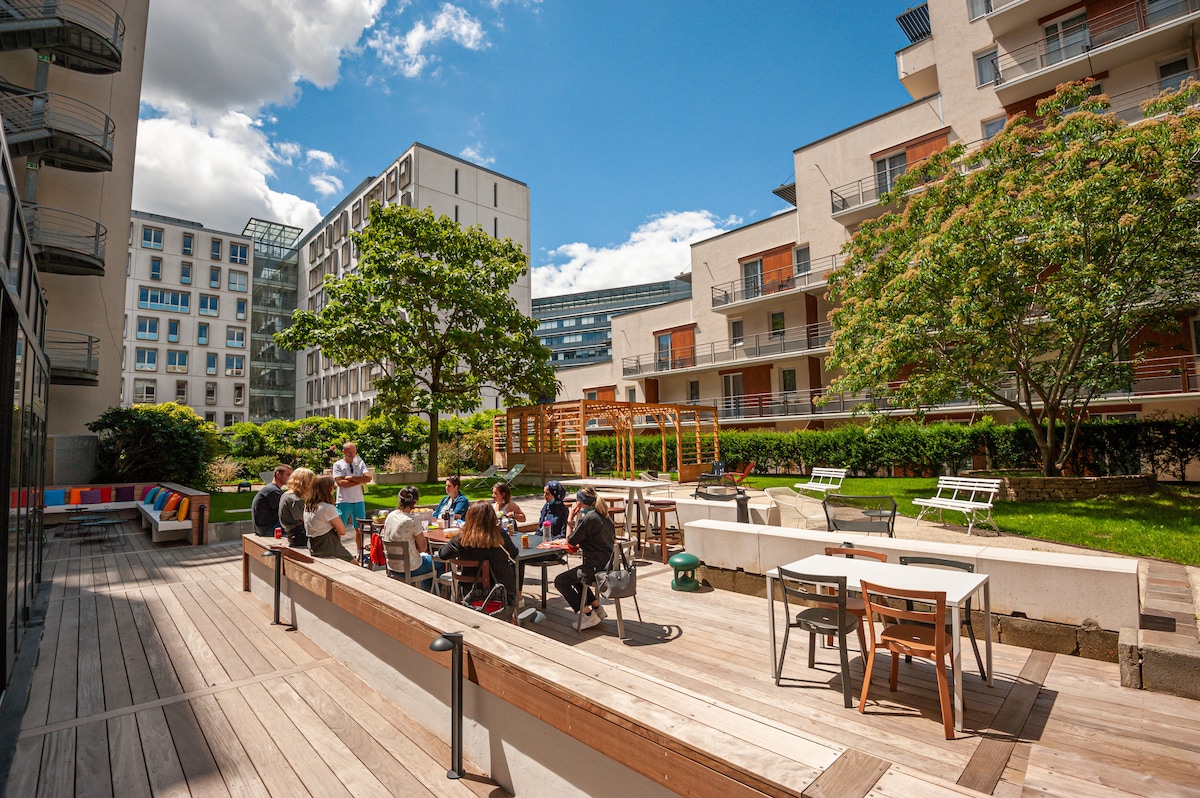 An outdoor communal area features a wooden deck with multiple seating arrangements, including tables and chairs. Residents are gathered, enjoying conversations under a clear blue sky. Lush greenery surrounds the space, contributing to a relaxed environment with a playground visible in the background.