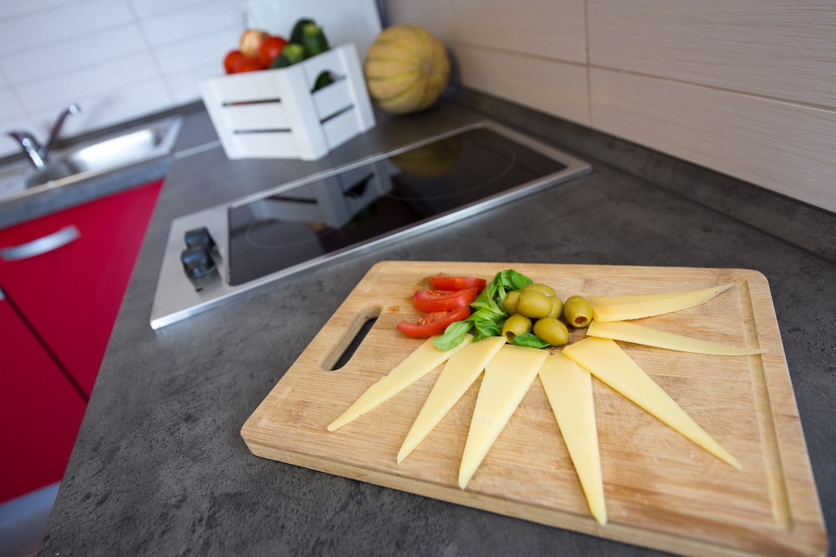 A wooden cutting board is displayed with sliced cheese arranged in a sunburst pattern, accompanied by green olives and tomato slices. A modern electric cooker is visible in the background, along with a basket of assorted vegetables resting on the counter.