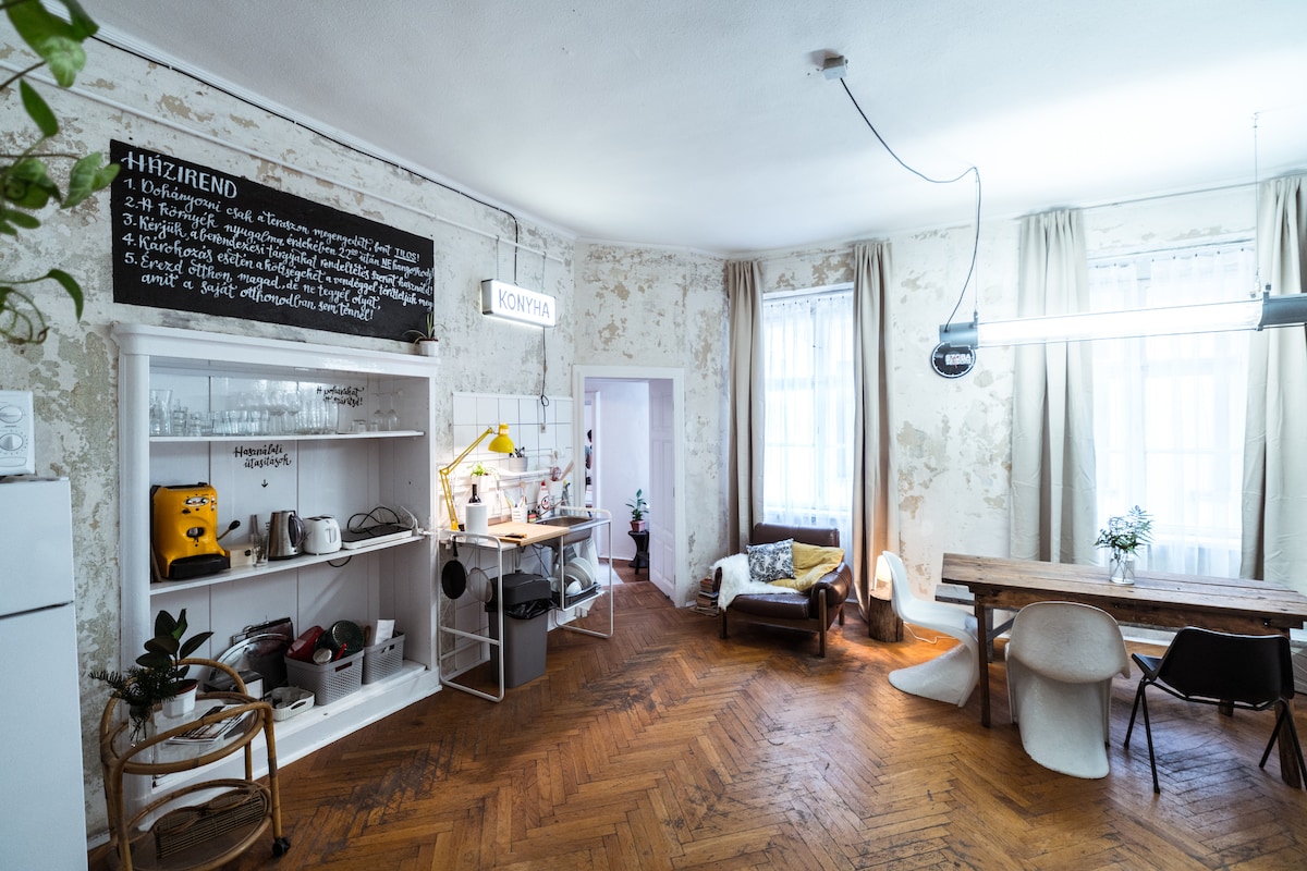 The living area features exposed walls that reflect the building's history. A wooden dining table is accompanied by light-colored chairs. A small kitchenette is equipped with basic appliances, while a cozy seating arrangement includes a brown leather sofa and a chair. Natural light filters through the large windows.