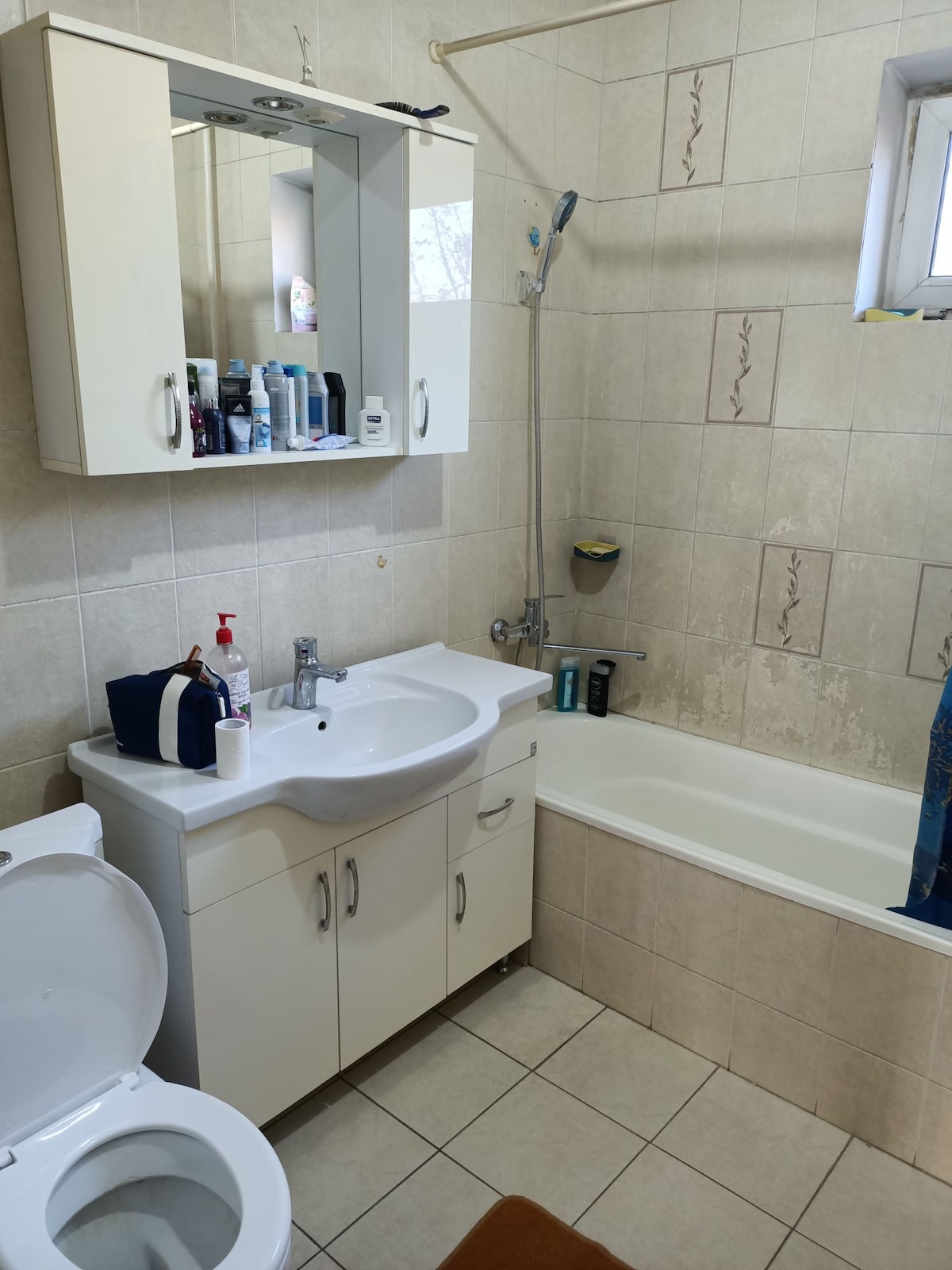 A light and neutral-colored bathroom features a white sink with a mirror above it and a combination bathtub and shower. Shelves hold toiletries, while a dark blue shower curtain adds a touch of color. Floor tiles are laid in a grid pattern.
